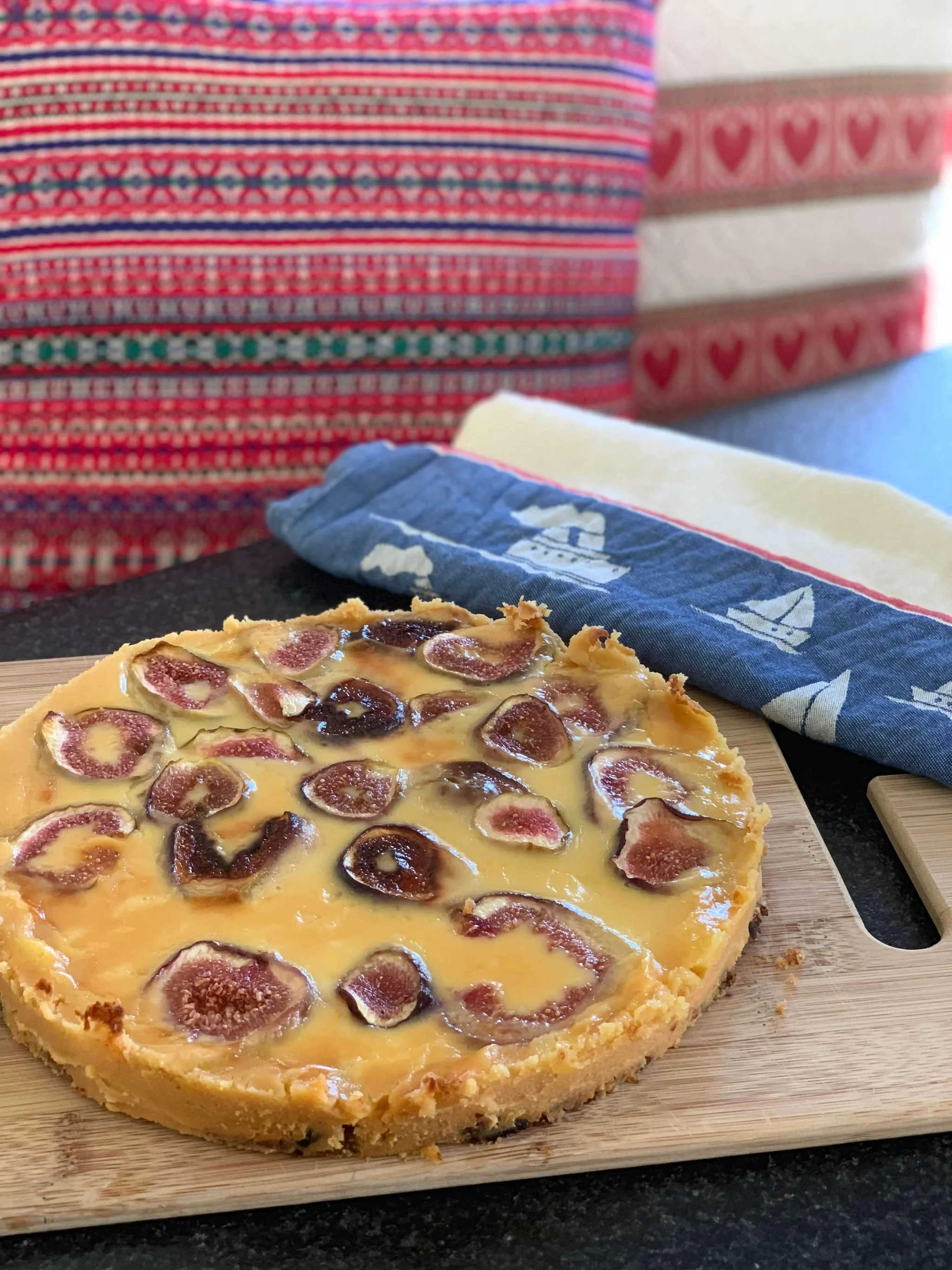 Round tart topped with sliced figs on a wooden board with colorful patterned cloths in the background