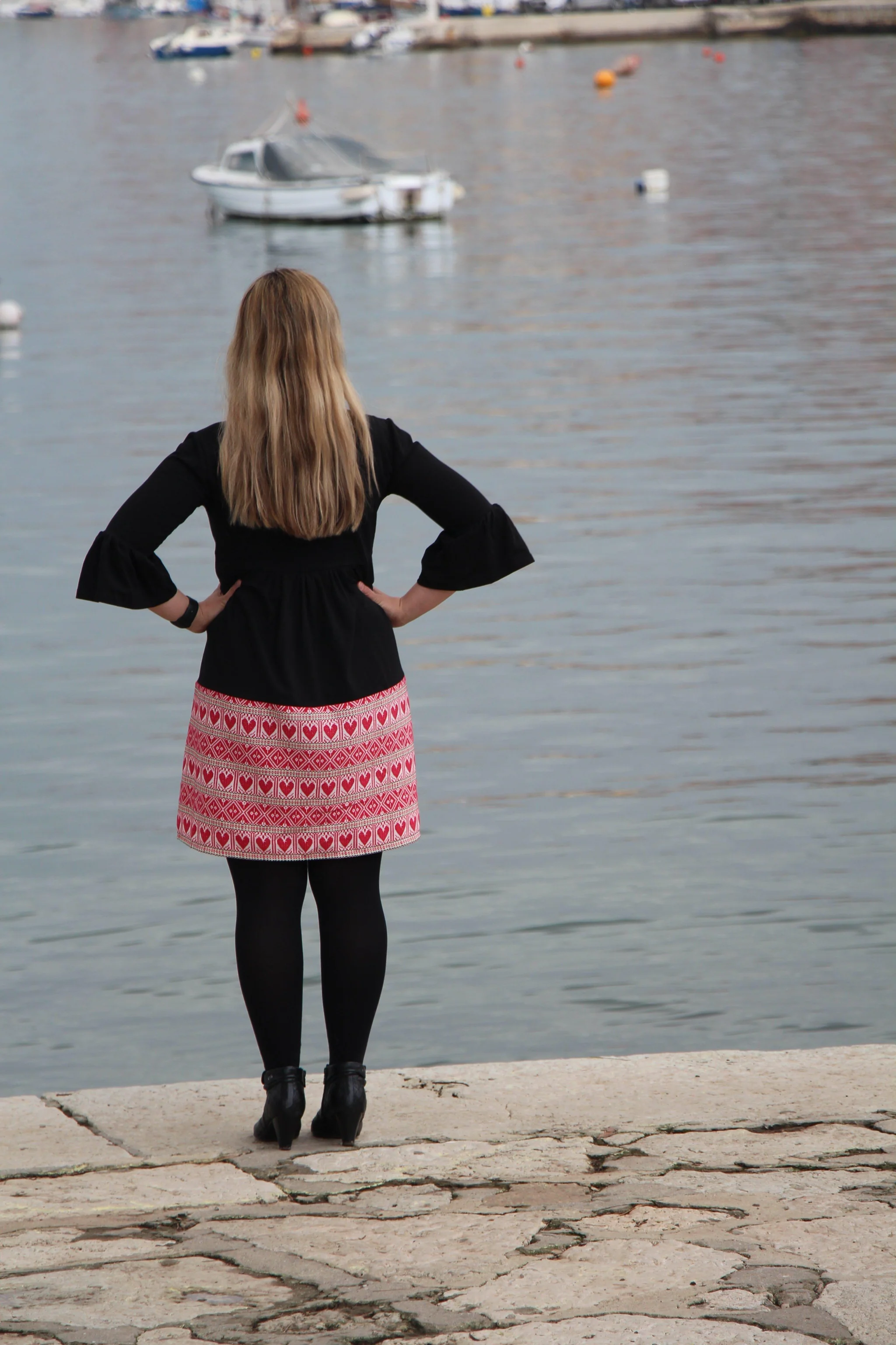 Woman viewed from behind facing a harbor with a boat, wearing a black dress with red heart-patterned hem